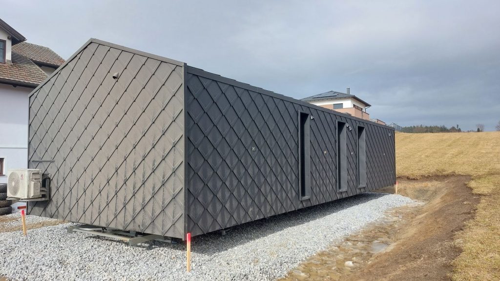Rückansicht des Modulhauses ATB 39 mit Rautenschindeln, schmalen Fenstern und Klimagerät, bewölkter Himmel.