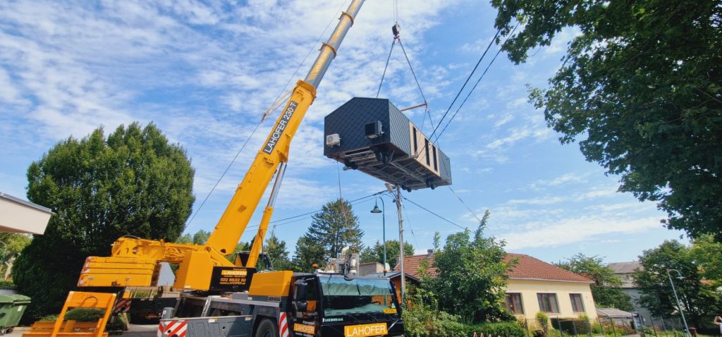 Schwebendes Premium-Modul mit Holzfront und dunklem Giebel am Kran über einem ruhigen Wohnviertel.