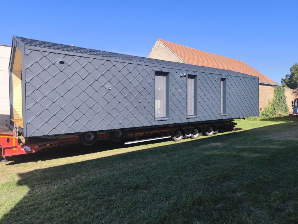 Lange Seitenansicht des Fertighauses ATB 53 mit Rautenfassade und drei hohen Fenstern im Sonnenlicht.
