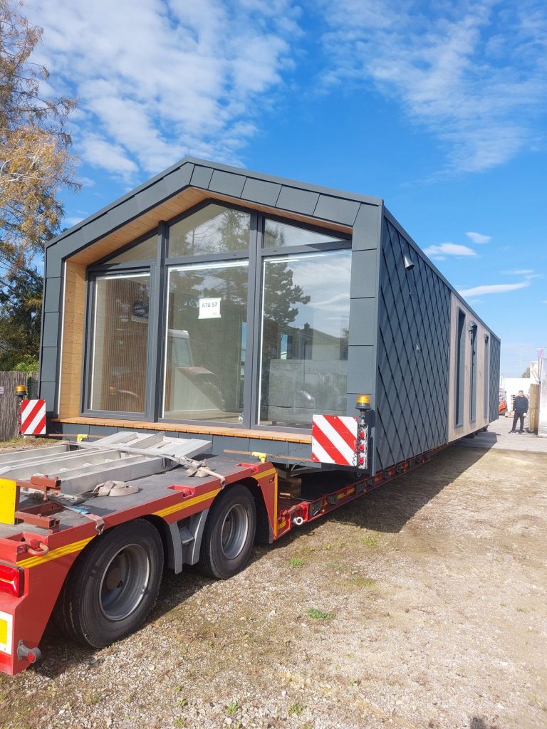 Verglaste Giebelfront des ATB 57 mit Panoramafenstern und Holzleibungen, transportiert auf rotem Tieflader.