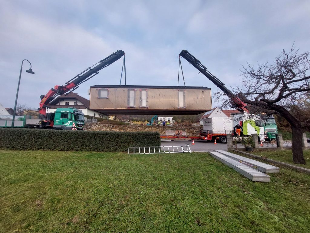 Zwei Ladekräne heben ein transportverkleidetes BOND Modulhaus über die Steinmauer auf das Grundstück.
