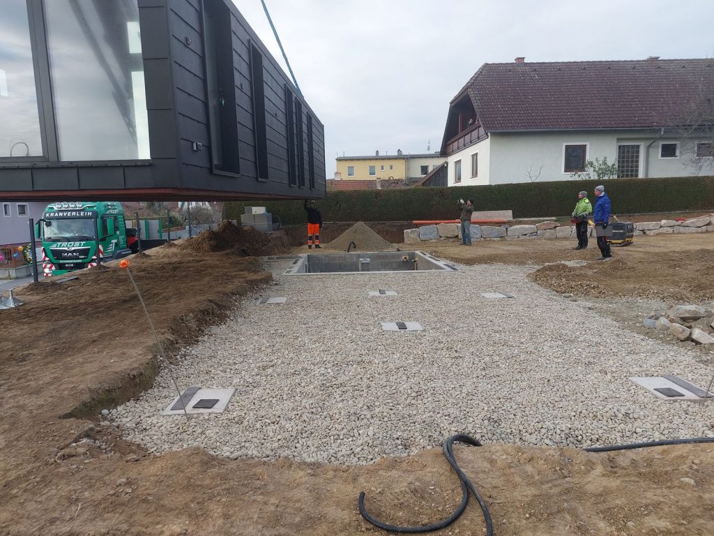 Schwarzes Fertighaus-Modul schwebt am Kran über dem vorbereiteten Kiesbett mit Punktfundamenten.