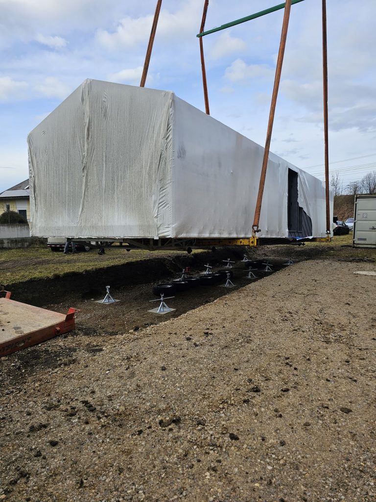 Folienverpacktes Modul mit Satteldachform schwebt am Kran über den vorbereiteten Fundamentstützen.