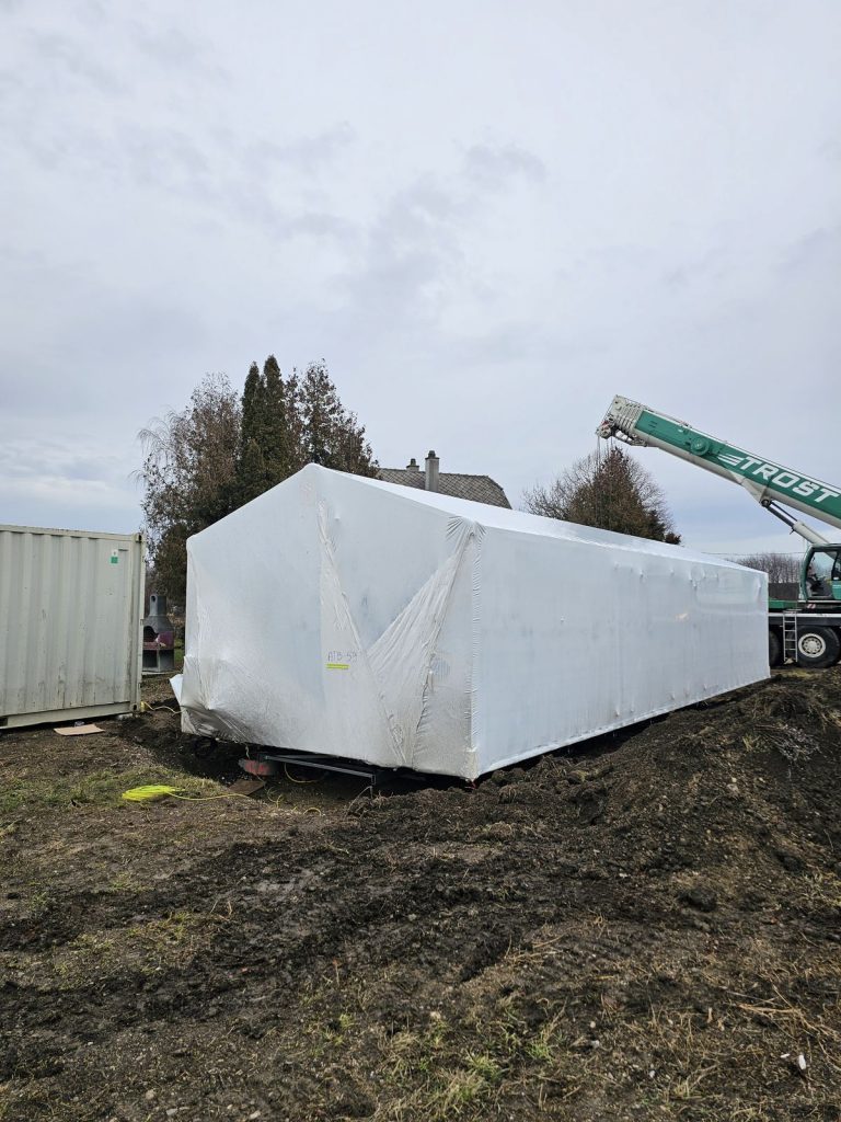 Mit ATB 59 beschriftetes, folienverpacktes Hausmodul vor dem grünen Mobilkran am Lieferort.