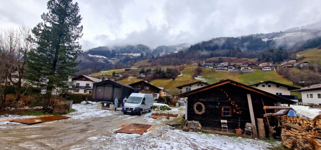 Das Modulhaus ATB 60 eingebettet in ein alpines Dorf mit Holzhütte, verschneiten Hängen und Nebel.