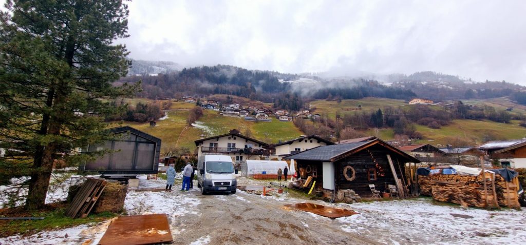 Weite Winterlandschaft mit dem ATB 60 links, Bergdorf, Gewächshaus und bewaldeten Hängen im Hintergrund.