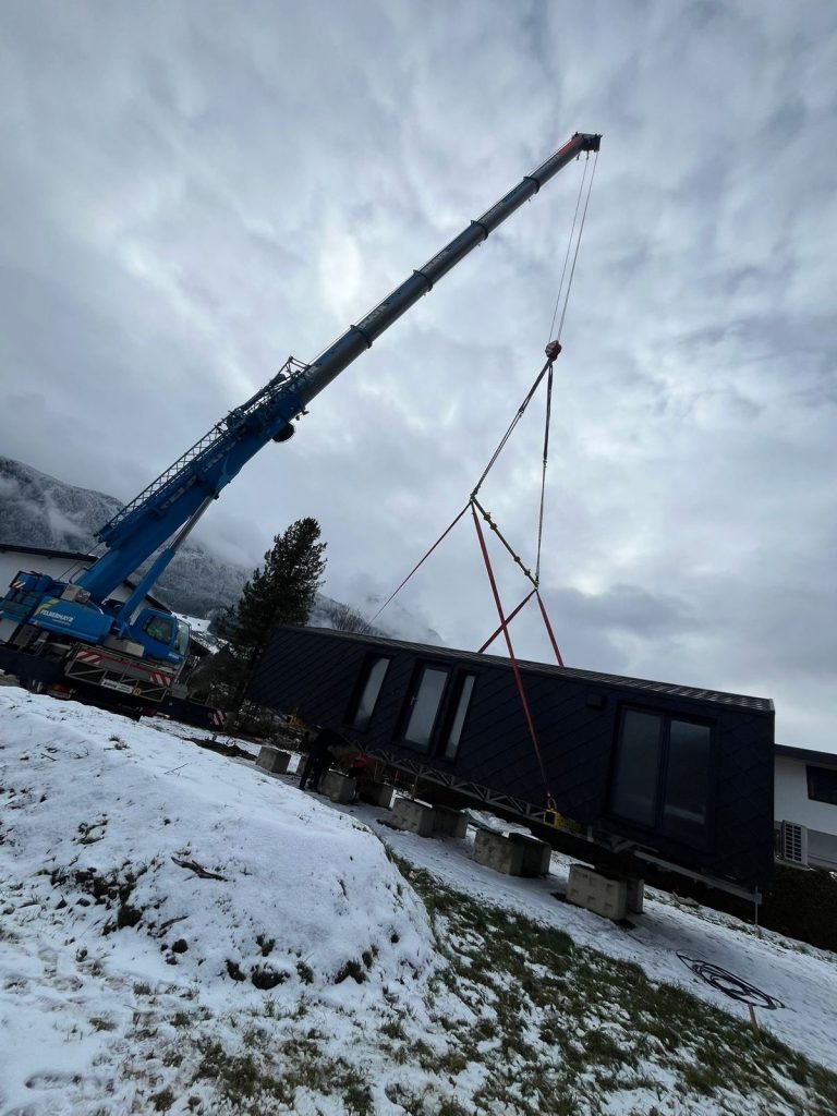 Der Kran hebt das Modulhaus ATB 60 mit roten Hebegurten sicher an seinen Platz am verschneiten Hang.