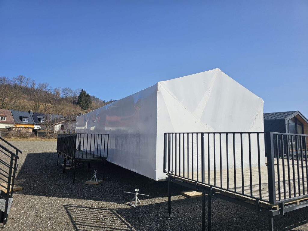 Transportfertiges BOND Modul in Schutzfolie neben fertigem Modulhaus mit Giebelfenster, blauer Himmel
