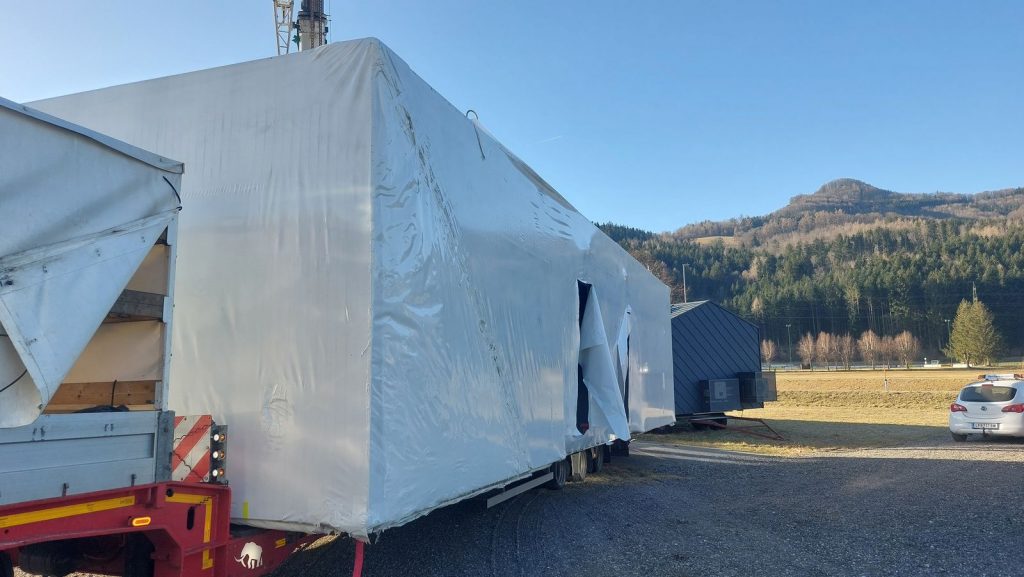 In weiße Schutzfolie verpacktes BOND-Modulhaus auf dem Tieflader vor bewaldeten Hügeln, bereit zur Anlieferung.