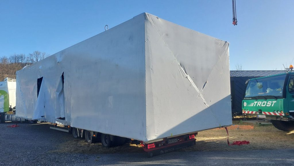 Rückansicht des folienverpackten Fertighaus-Moduls auf dem Transportanhänger neben einem grünen Kranwagen.
