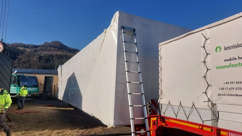 Folienverpacktes Wohnmodul mit angelehnter Teleskopleiter während der Anlieferung, daneben Kranwagen und Team.