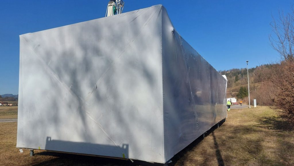 Folienverpacktes Modulhaus unter dem Kranausleger auf dem Grundstück, ein Monteur steht zur Sicherung daneben.