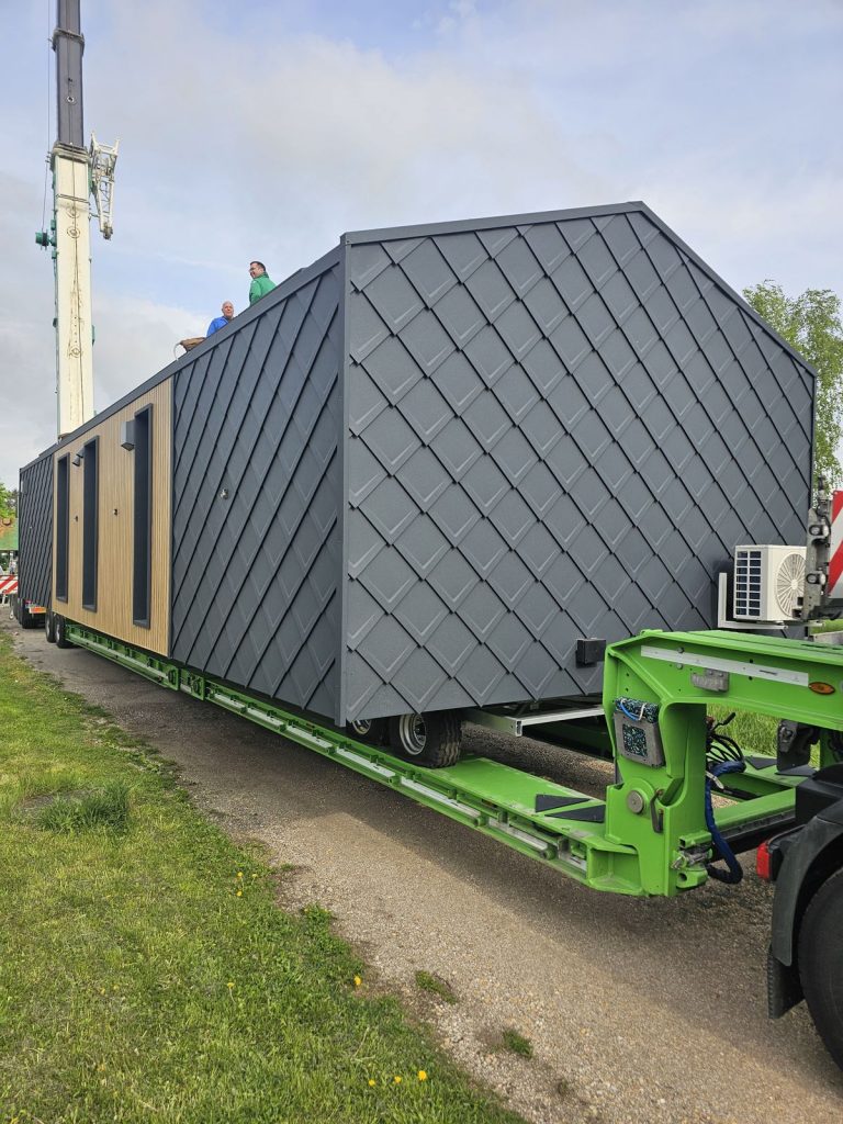 Anthrazitgraues BOND Modulhaus mit Rautenschindel-Fassade auf grünem Tieflader vor dem Transport.