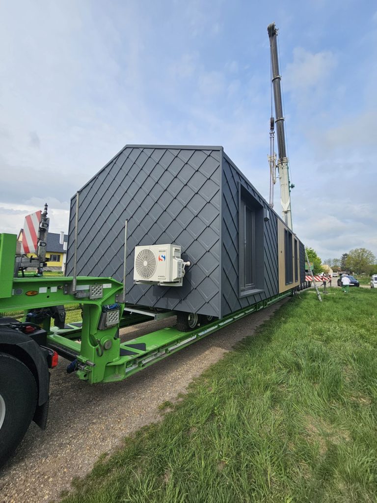 Anthrazitgraue Rautenschindel-Fassade des ATB 64 mit Klimagerät, angeliefert per Kran auf grünem Tieflader.