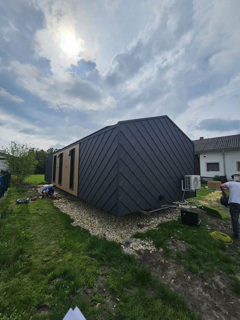 Aufgestelltes BOND Ganzjahreshaus mit anthrazitfarbener Metallfassade auf Kiesbett während der Montage im Garten.