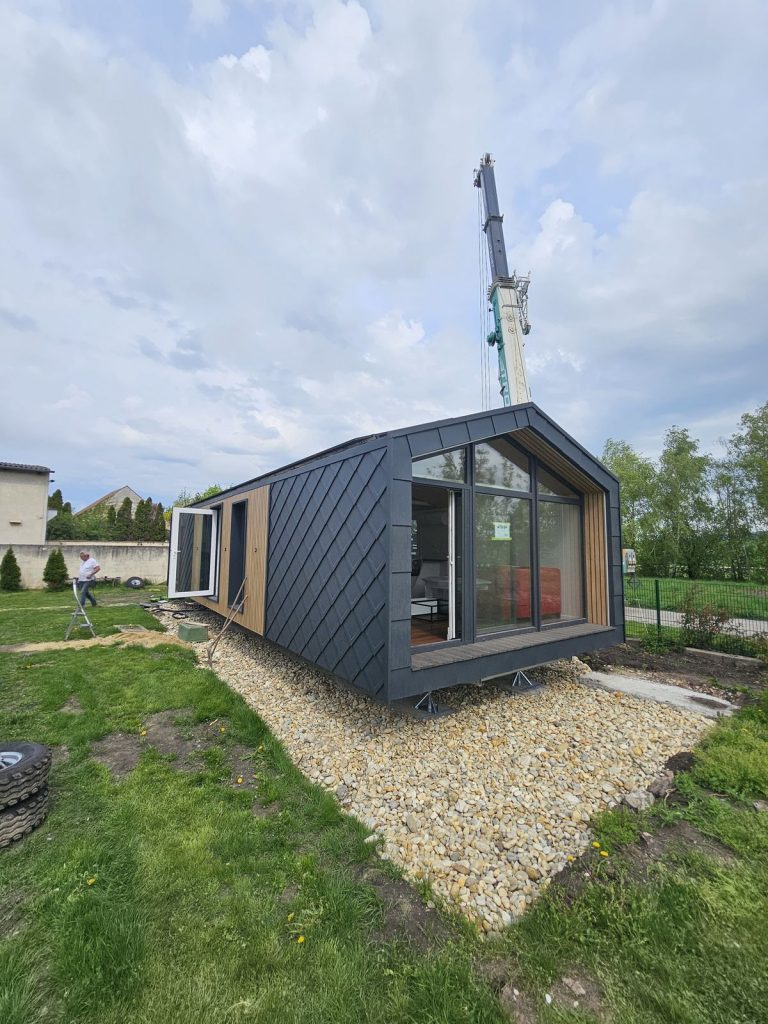 Schlüsselfertiges ATB 64 mit verglaster Giebelfront, offener Tür und Holzterrasse auf dem Grundstück.