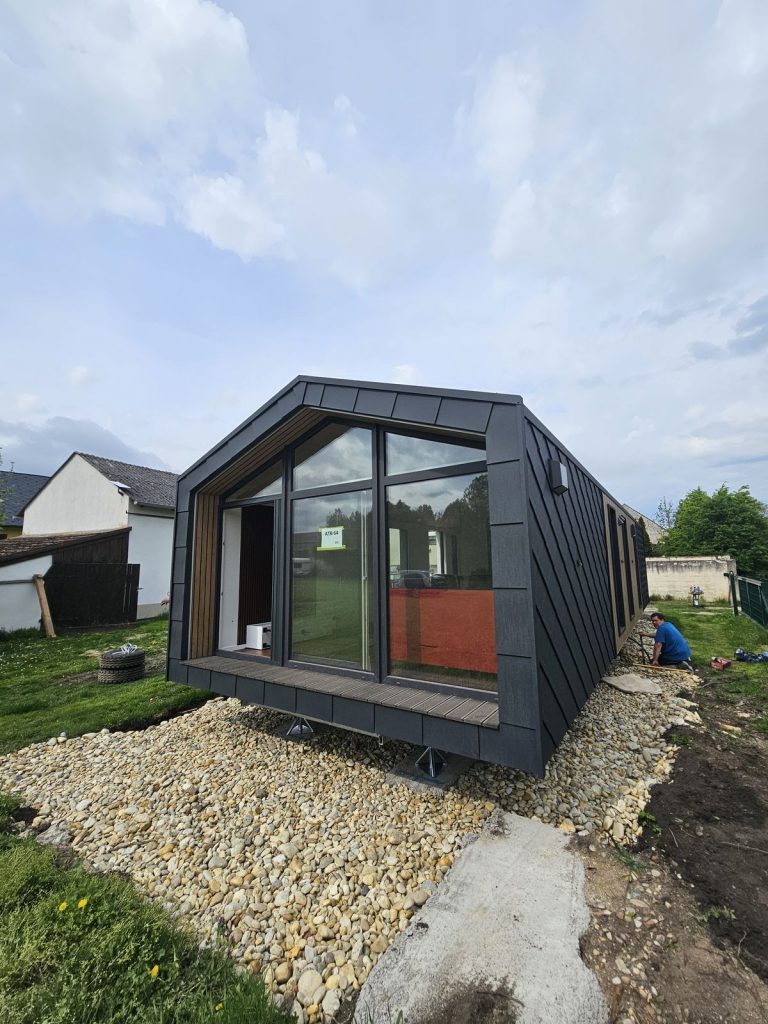 Fertig aufgestelltes Premium-Modulhaus ATB 64 mit Panorama-Giebelfront, Holzterrasse und Rautenfassade im Garten.