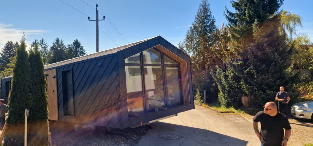 Giebelseite des Modulhauses mit raumhoher Panoramaverglasung und warmem Holz, angeliefert auf dem Anhänger
