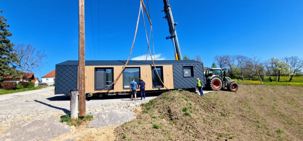 Kran hebt das schlüsselfertige Modul mit Holzfront und Fensterband über die vorbereitete Baustelle