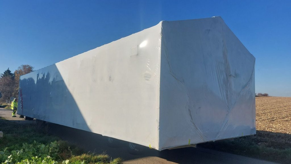 In Schutzfolie verpacktes Fertigteilhaus-Modul auf dem Transportanhänger neben einem gepflügten Acker.
