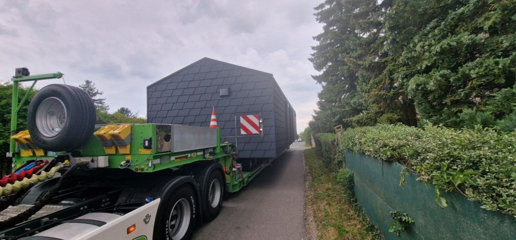 Schlüsselfertiges Modulhaus auf grünem Tieflader auf schmalem Weg zwischen Hecken bei der Anlieferung.