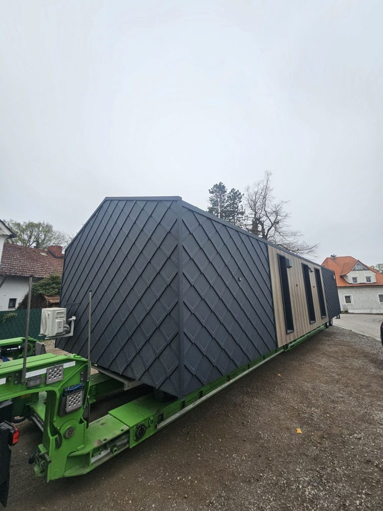 Längsseite des ATB 71 mit Rautenschindeln und warmer Holzfassade samt drei schmalen Fenstern vor Ortskulisse.