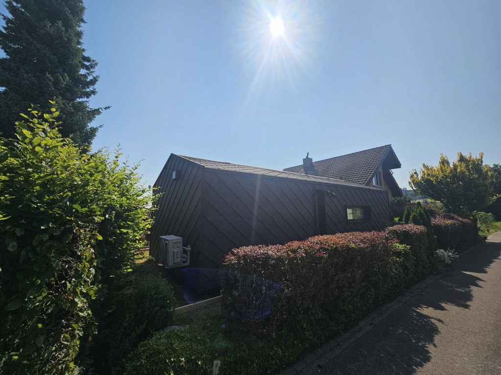 Dunkles mobiles Eigenheim hinter dichter Hecke mit asymmetrischer Dachform bei sonnigem Gegenlicht.