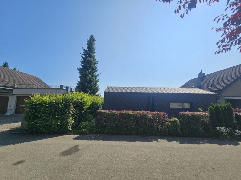 Straßenansicht des dunklen Premium-Modulhauses hinter der Hecke, eingebettet zwischen Nachbarhäusern.