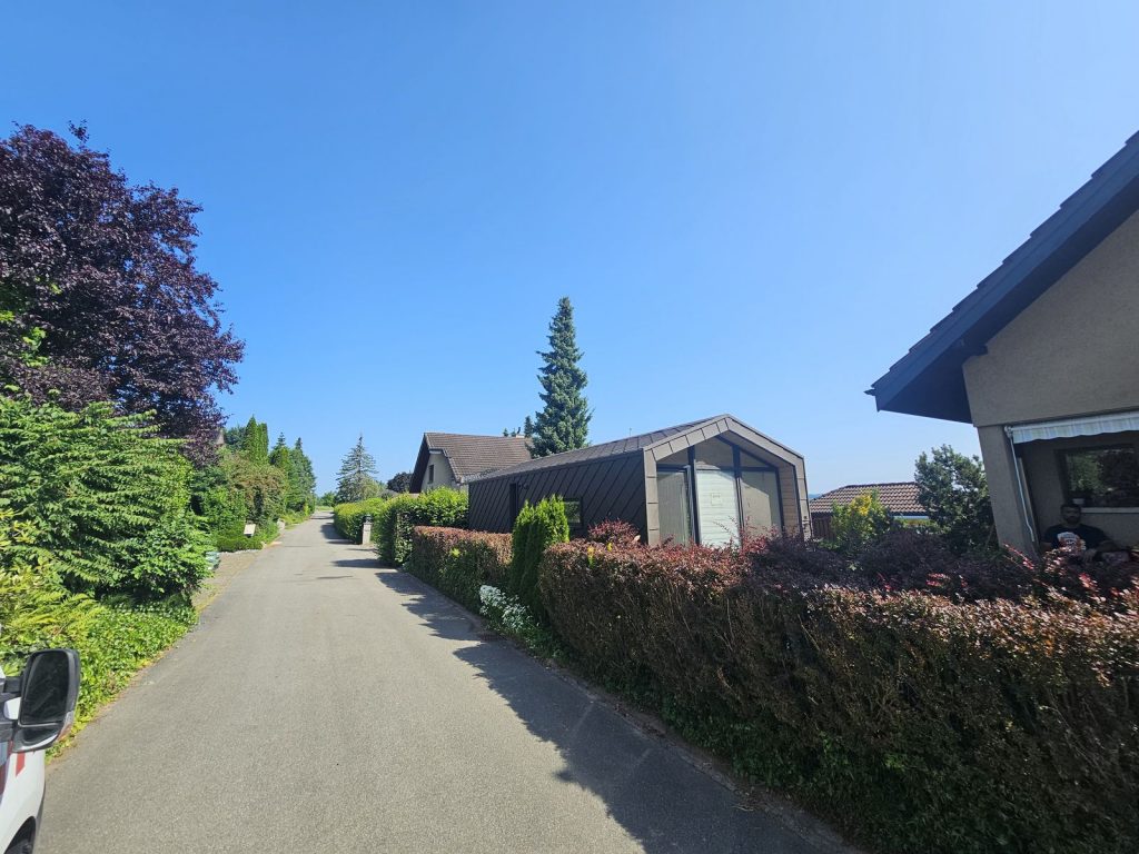 Blick entlang der Anliegerstraße auf das dunkel verkleidete Modul mit verglaster Giebelseite hinter der Hecke.