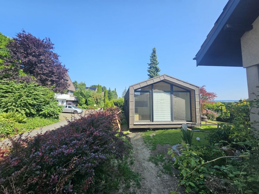 Verglaste Giebelseite mit Terrasse im Garten, dahinter Nadelbaum und weiter Ausblick über die Landschaft.