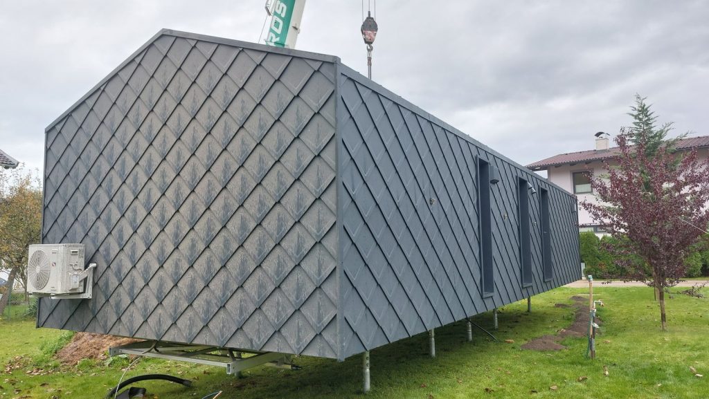 BOND Modulhaus mit anthrazitfarbener Rautenschindel-Fassade und Wärmepumpe, aufgestellt auf Stützen im Garten.