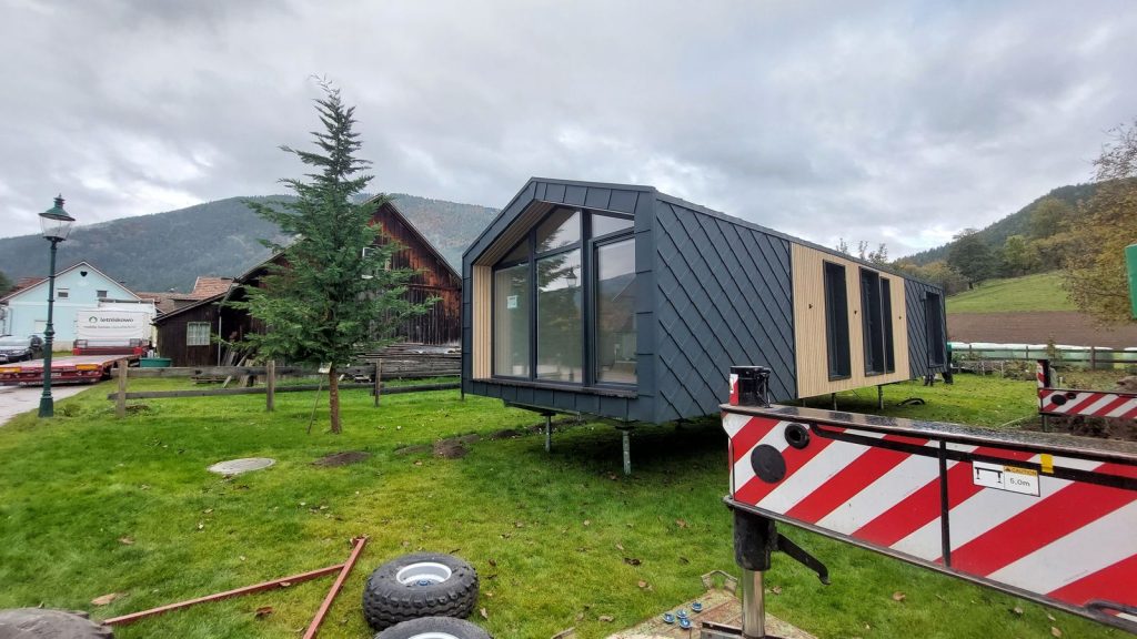 Modulhaus in Schrägansicht mit Giebelfenster und Metall-Rautenschindeln, Kranarm im Vordergrund.