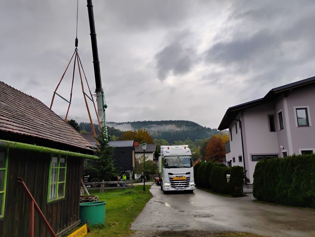 Anlieferung des Modulhauses per Sattelzug durch eine Ortsdurchfahrt unter bewölktem Himmel.