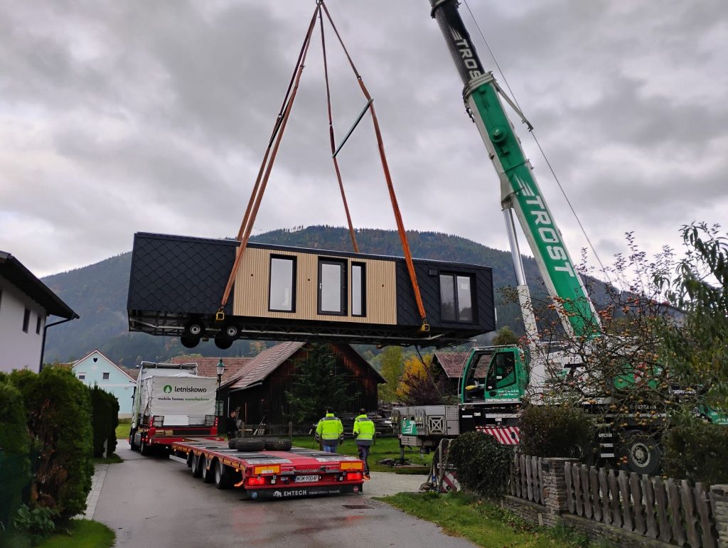 Fertiggestelltes Modul mit Lärchen-Holzfassade schwebt am Kran über dem Tieflader.