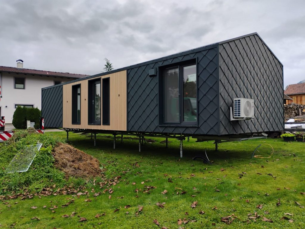 Seitenansicht des vollmöblierten Modulhauses mit Holzlattung, Eingangstür und Rautenschindel-Fassade.