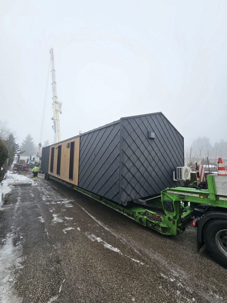 Das schlüsselfertige Modulhaus ATB 76 in voller Länge auf dem Tieflader mit Rautenfassade und Holzstirnseite.