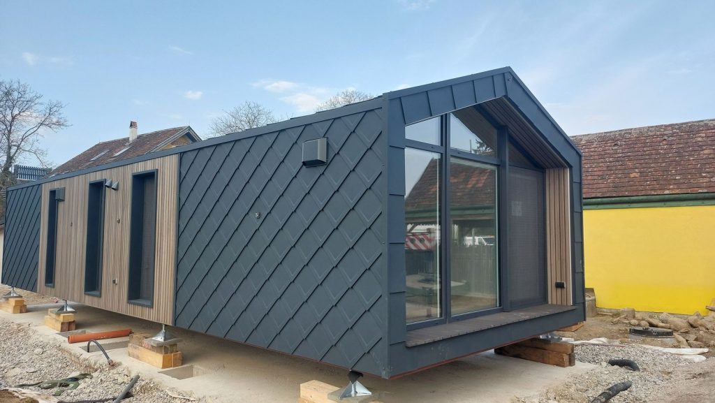 Längsseite des ATB 77 mit dunkler Rautenfassade, Holzverkleidung und Fenstern, dahinter ein Bauernhaus