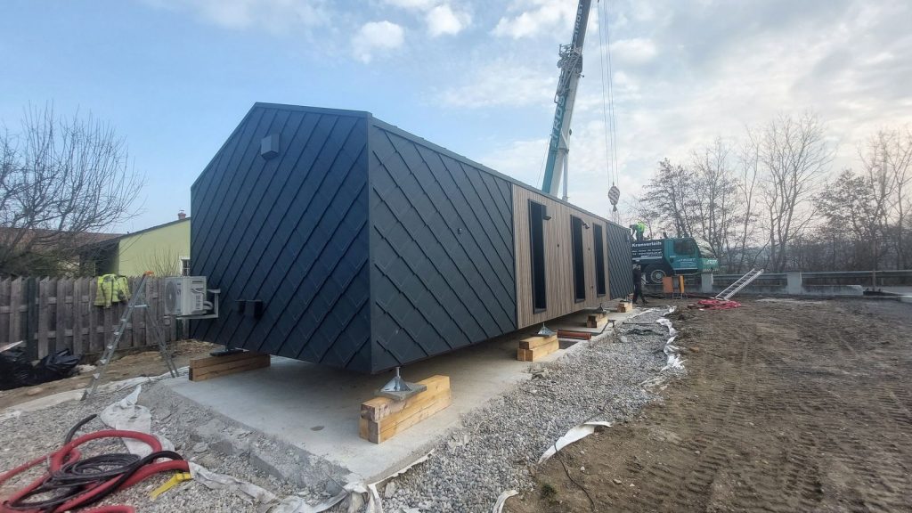 Kran setzt das Fertigteilhaus mit anthraziter Metallfassade auf die vorbereitete Betonplatte