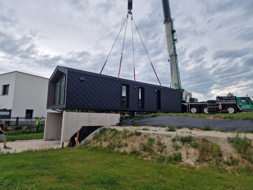 Kran hebt ein dunkles BOND Fertighaus-Modul mit Giebelverglasung über den Betonsockel