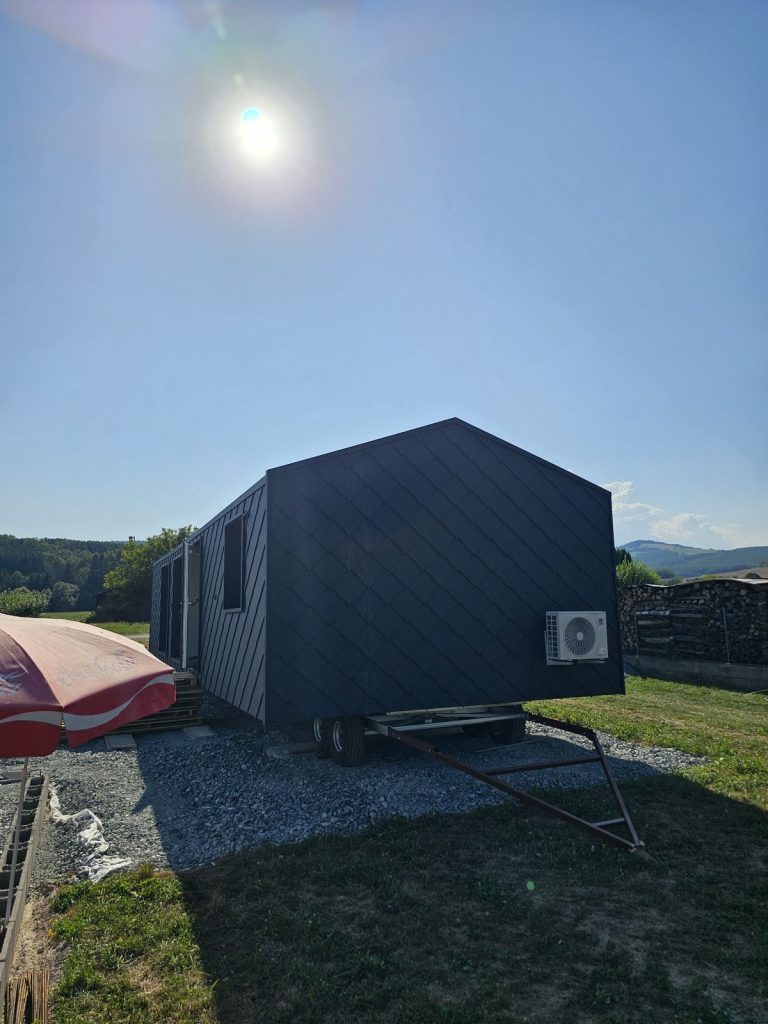 Giebelseite des dunklen Modulhauses mit Rautenfassade und Außeneinheit der Wärmepumpe im sommerlichen Gegenlicht.