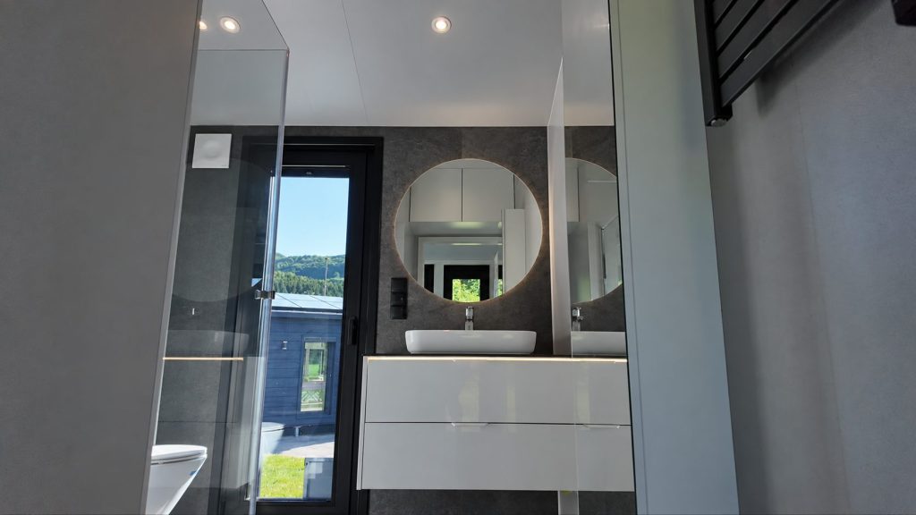 Bad im Modulhaus Diamond +Plus ATB-38 mit Waschtisch, Spiegel, Glasdusche und grauer Wandverkleidung.