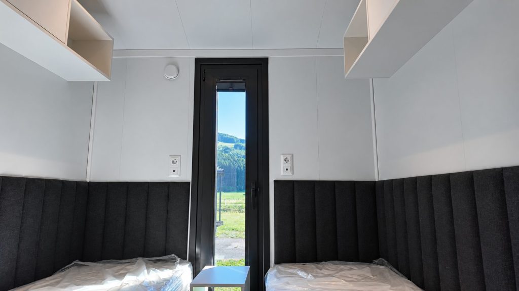 Gästezimmer im Modulhaus Diamond +Plus ATB-38 mit gepolsterter Wandverkleidung und schmalem Fenster.