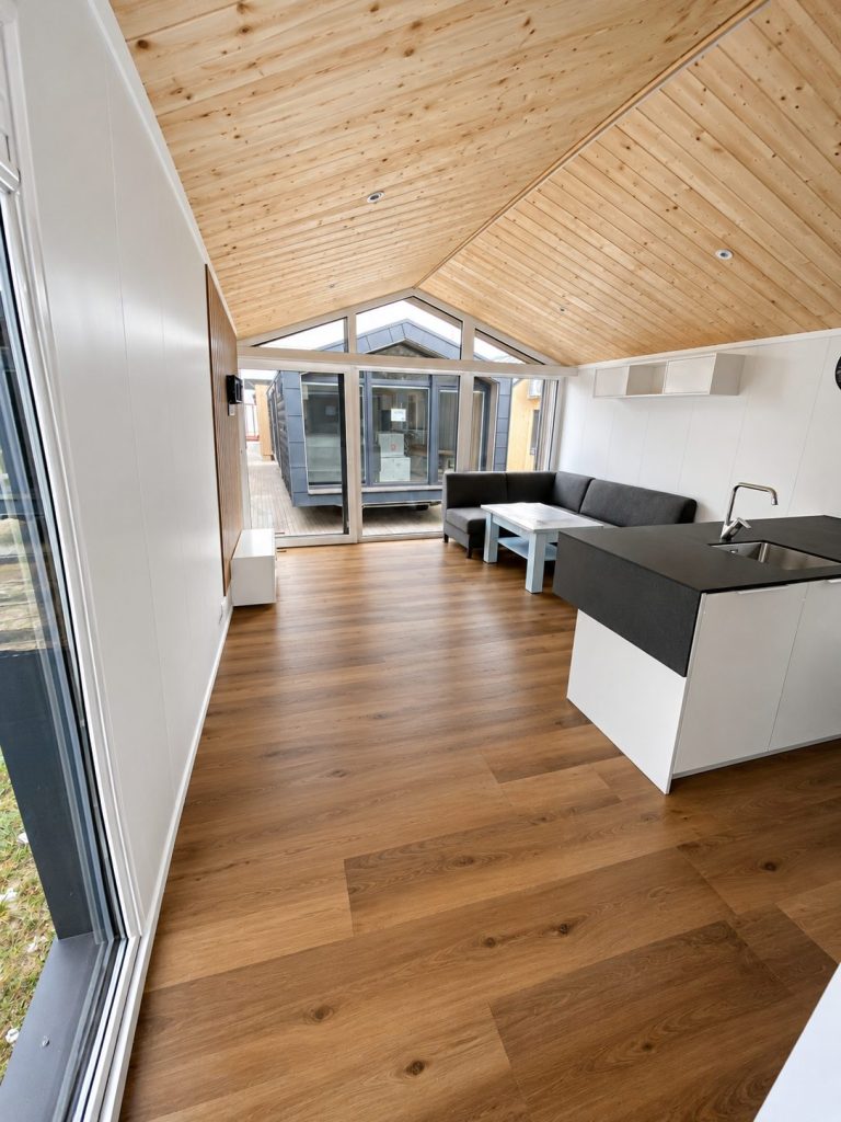 Wohnbereich im Modulhaus Diamond +Plus Deluxe ATB-62 mit Holzdecke, Giebelverglasung, Sofa und Kücheninsel.