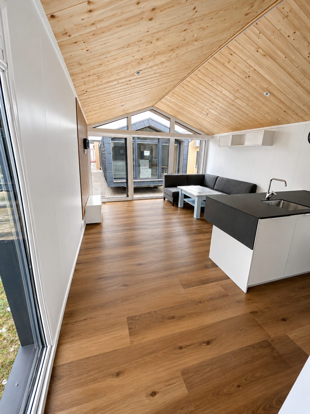 Wohnbereich im Modulhaus Diamond +Plus Deluxe ATB-62 mit Holzdecke, Giebelverglasung, Sofa und Kücheninsel.